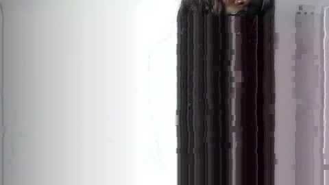 Video of a person with long, dark hair, partially obscured by a tall, dark curtain, standing against a plain, white wall in an indoor setting. The image is blurred and pixelated.