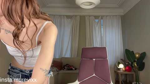 Video of a woman with long brown hair wearing a white tank top, leaning over a purple chair in a modern living room with large windows and a cactus plant.