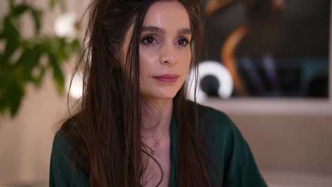 Video of a young woman with long, dark hair, wearing a green blouse, against a blurred indoor background with green plants and modern decor.