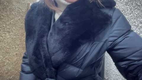 Video of a person wearing a black, puffy winter coat with a thick, plush black fur collar, standing against a textured, beige stone wall.