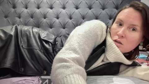 Video of a fair-skinned woman with brown hair, lying on a bed with a gray, tufted leather headboard. She wears a cream sweater and black leather overalls, appearing casual and relaxed.