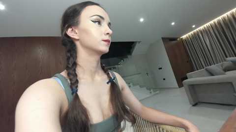 Video of a slender, fair-skinned woman with long brown hair in twin braids, wearing a green tank top, captured from a side angle in a modern, minimalist living room with wooden walls and a gray sofa.