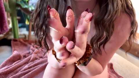 Video of a young woman with long brown hair, fair skin, and maroon nail polish, showcasing her hands with claws, sitting on a pink blanket indoors.