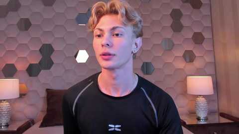 Video of a young, fit, fair-skinned man with wavy blonde hair, wearing a black athletic shirt with white accents, standing in a modern, hexagonal-patterned bedroom with two bedside lamps.