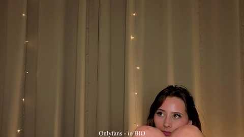 Video of a nude woman with long dark hair, pressing her face against sheer beige curtains, illuminated by soft, warm light.