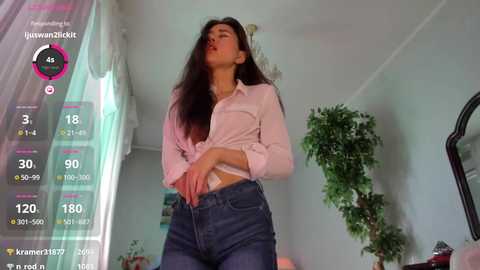 Video of an East Asian woman in a white button-up shirt and blue jeans, standing in a brightly lit room with a large potted plant, looking at her phone.