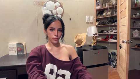 Video of a woman with dark hair in a messy bun, wearing a maroon off-shoulder sweatshirt with \"88\" in white, standing in a cluttered closet with shoes, hats, and a lamp.