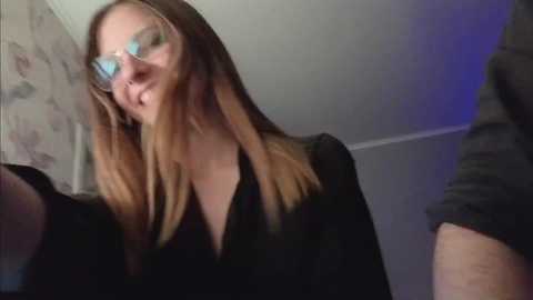 Video of a young woman with straight, light brown hair, wearing clear aviator glasses and a black top, smiling, with a textured floral wallpaper in the background.