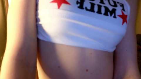 Video of a pale-skinned person wearing a white crop top with \"GOLD STAR\" printed in black and red stars, revealing a flat stomach and light moles. Background is out of focus.