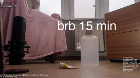 A video shows a person sitting on a pink towel-covered wheelchair, with a transparent bottle and a white tube on the wooden floor. The background features a radiator and a window with blinds.