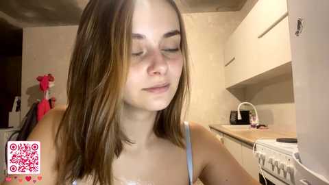 Video of a young woman with long brown hair, fair skin, and closed eyes, wearing a light blue spaghetti-strap top, standing in a small kitchen with white cabinets and a white oven.