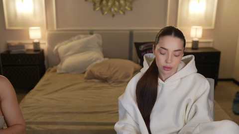 Video of a serene woman in a white robe, eyes closed, on a bed with beige sheets and pillows, surrounded by dimly lit nightstands with lamps, in a cozy, minimalist bedroom.