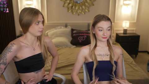 Video of two young women with long hair, one wearing a black tube top, the other a navy blue one-piece. They're in a bedroom with a bed, wall art, and lamps.
