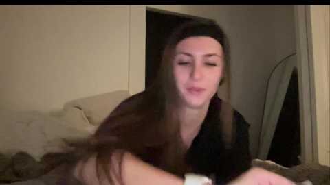 A blurry video of a woman with long brown hair lying on a bed, wearing a black top, in a dimly lit room with beige walls and a mirror.