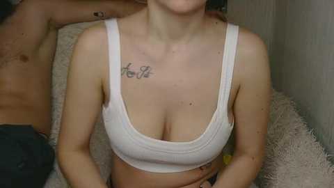 Video of a light-skinned woman with a tattoo on her collarbone, wearing a white sports bra, seated on a fluffy cream-colored sofa.