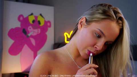 Video of a topless, fair-skinned blonde woman with large breasts, applying lipstick in a dimly lit room. Background features a pink cartoon character and a glowing neon sign.