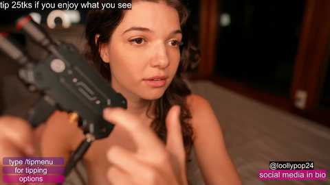 Video of a topless young woman with curly brown hair, holding a camera. She appears confused, with a wooden door in the background. Text overlays suggest a \"tip\" to watch the video.