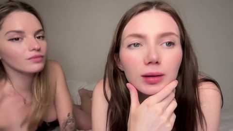 Video of two young, fair-skinned women with long hair, one topless, the other partially visible, both with light makeup, lying on a bed with neutral bedding, in a soft-lit room.