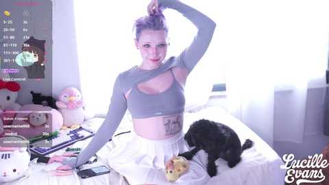 Video of a young, pale-skinned woman with purple hair, wearing a gray crop top, white skirt, and black cat. She sits on a bed, surrounded by plush toys and a keyboard.
