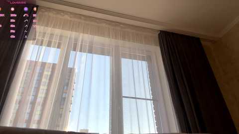 Video of a window with sheer white curtains and dark curtains pulled to the side, showing a clear blue sky. Building and street view visible through the window.