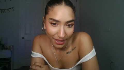 Video of a young, light-skinned woman with medium-length dark hair, wearing a white off-shoulder top, silver hoop earrings, and a delicate necklace. She has a small tattoo on her shoulder. Background is a dimly lit room with a closed door and a white wall.
