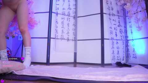 A video of a nude, light-skinned woman with a slender build, wearing white socks, standing in front of a Japanese room divider with cherry blossom prints.