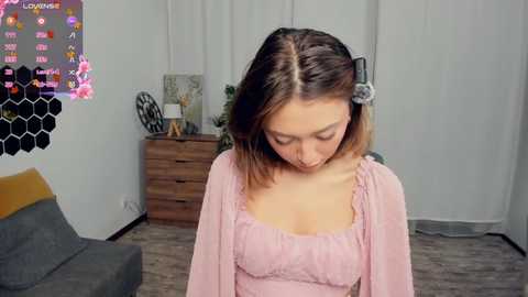 Video of a young woman with shoulder-length brown hair, wearing a pink blouse, standing in a modern living room with wooden furniture, a hexagonal artwork, and a gray couch.