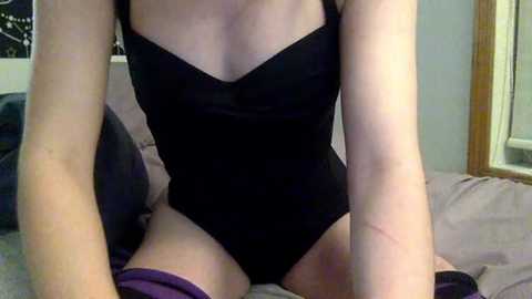 A video of a light-skinned woman in a black, snug-fitting, halter-neck bodysuit, kneeling on a bed with light gray sheets, showing her arms and thighs.