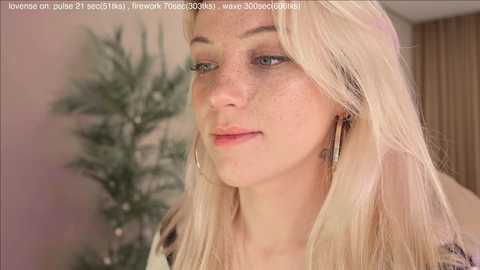 A close-up video of a fair-skinned woman with long, straight blonde hair, wearing large hoop earrings, and a light-colored top. She has freckles and blue eyes, with a soft, natural makeup look. The background features a blurred, green Christmas tree and a beige wall.