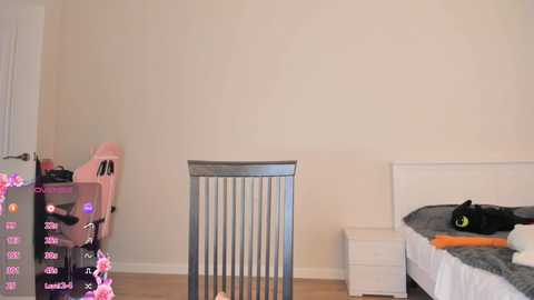 Video of a minimalist bedroom with a white bed, gray blanket, black cat, and a metal chair. A pink high chair is positioned against a cream-colored wall.