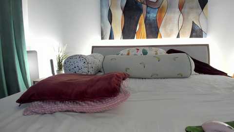Video of a modern bedroom with a white bed, a teal curtain, a colorful abstract painting, and a decorative pillow on a white pillowcase.