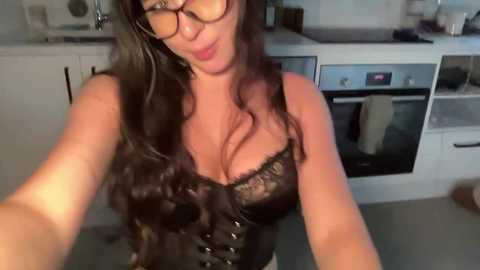 Video of a woman with long, wavy dark hair, wearing glasses and a black lace corset, standing in a modern kitchen.