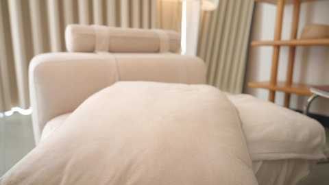 Video of a serene spa room with a massage table covered in white sheets, beige curtains, and a wooden shelf in the background.