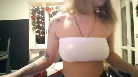 A video of a light-skinned woman with shoulder-length blonde hair, wearing a white bandeau top and black shorts, with tattoos on her arms. She stands indoors near a window adorned with Christmas decorations, holding a camera.