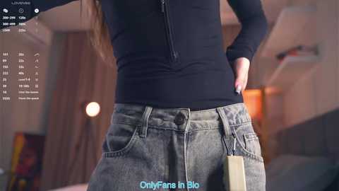 Video of a woman in a black long-sleeve top and worn-out jeans, holding a beige handbag, standing indoors with a blurred background. Text reads \"OnlyFans in Blue.\