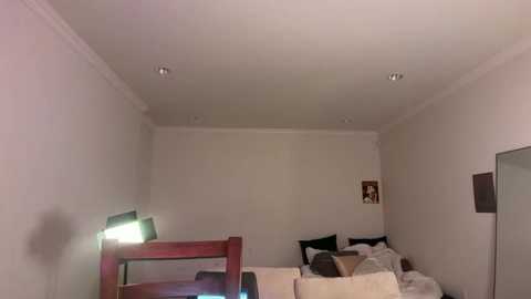 Video of a minimalist, dimly lit bedroom with white walls, a single bed, and a small nightstand with a lamp. The bed is unmade, and a framed picture hangs on the wall.