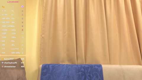 A video of a cozy living room with a beige curtain, a blue blanket on a cream sofa, and a yellow wall with a digital clock.