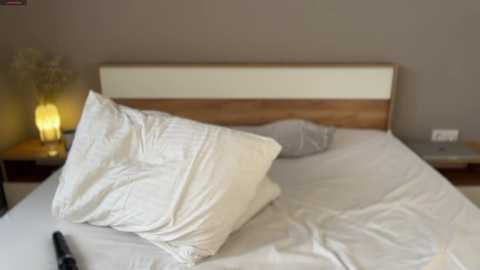 Video of a neatly made bed with white pillows and sheets, against a beige wall, featuring a wooden headboard and bedside table with a yellow lamp.