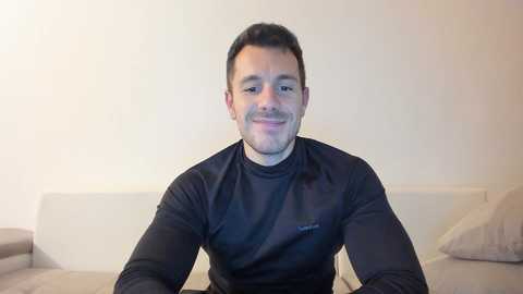 Video of a smiling, fit man with short dark hair, wearing a black long-sleeve shirt, seated on a beige couch in a minimalist, off-white room.