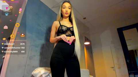 Video of a slim, light-skinned woman with long, straight blonde hair, wearing a black lace bralette and high-waisted black pants, standing in a modern bedroom with a mirrored wall, heart-shaped nails, and a live stream overlay.