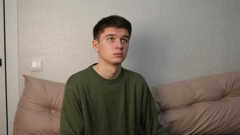 Video of a young man with short brown hair, wearing a green shirt, sitting on a beige tufted sofa against a light grey wall.