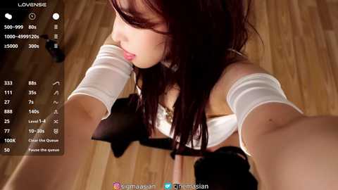 A video of an Asian woman with long brown hair, wearing a white off-shoulder top, taking a selfie indoors on a wooden floor.