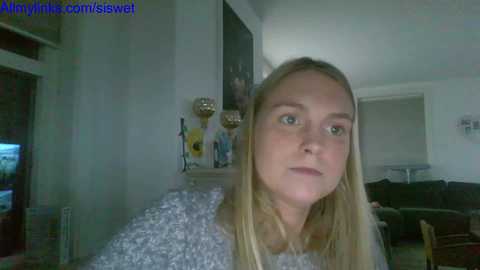 A video of a blonde woman with fair skin, wearing a textured sweater, looking pensive in a dimly lit living room.