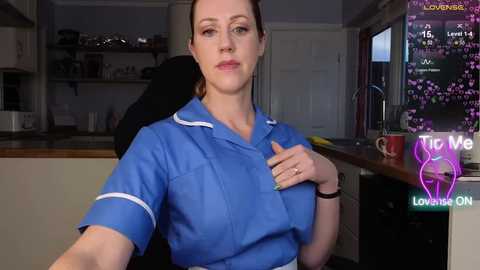 Video of a woman with fair skin, brown hair, and blue eyes, wearing a blue shirt, seated at a kitchen counter. The background shows a modern kitchen with cabinets and a window.