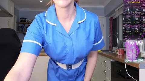 Video of a woman in a blue nurse's uniform with white trim, standing in a modern kitchen. The background features white cabinets, a pink mug, and a colorful wall calendar.