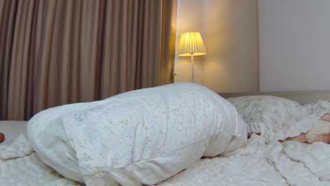 Video of a person lying in bed, covered by a white, patterned comforter. The room features beige curtains and a bedside lamp with a pleated shade, casting a warm glow.