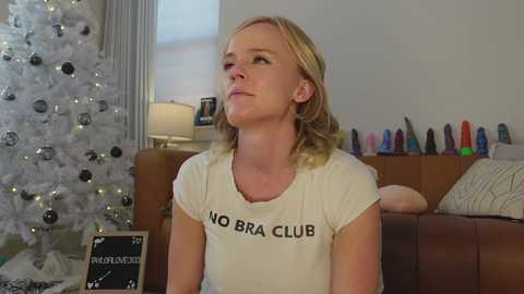 Video of a blonde woman in a beige \"NO BRA CLUB\" T-shirt, sitting on a brown couch, looking contemplative. Background features a white Christmas tree and a shelf of colorful sex toys.