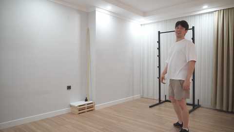 A video of a white man with short brown hair and glasses, wearing a white T-shirt and gray shorts, standing in a minimalist, well-lit gym with wooden floors and white walls.