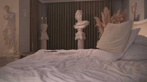 Video of a plush, white-sheeted bed with beige pillows and a gold-framed painting of a nude woman, set against a dark green curtain backdrop.