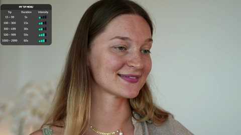 Video of a young woman with light skin and long, straight brown hair, smiling, wearing a gray top and gold necklace, against a blurred background.